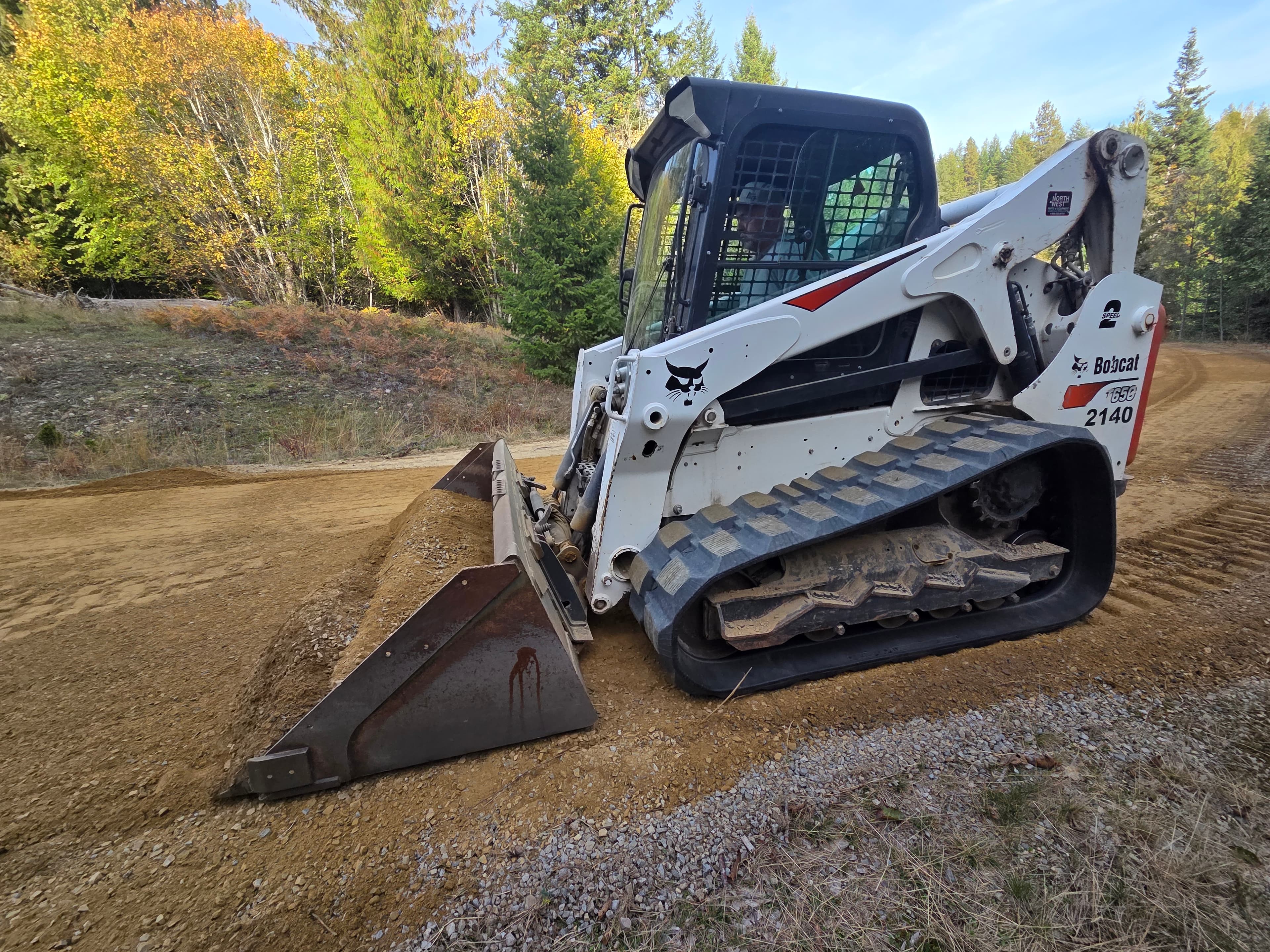 White Bobcat compact track loader levels a dirt road in a forest setting with autumn foliage.