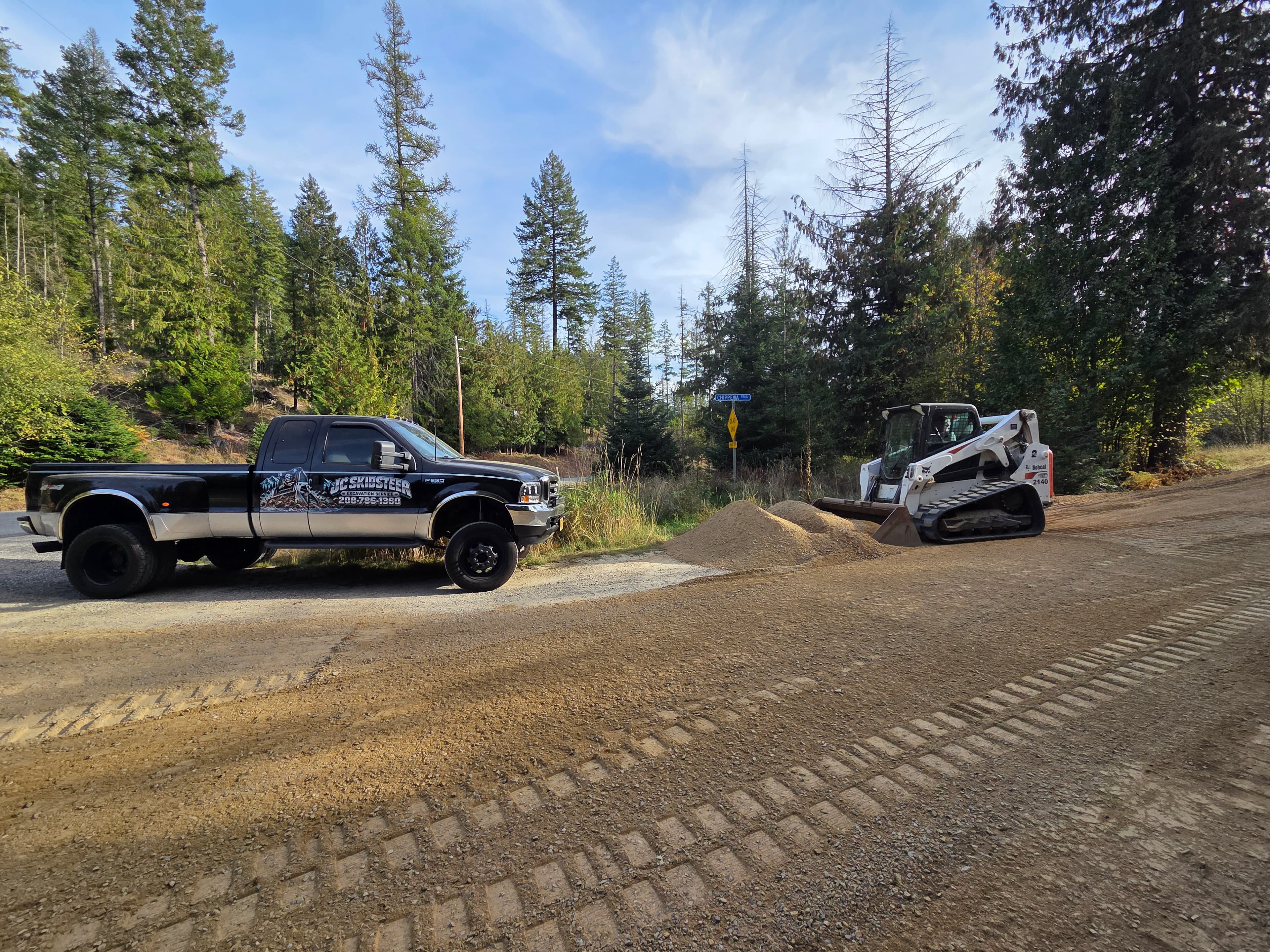 Black Ford truck and white Bobcat skid steer on a dirt road surrounded by pine trees.