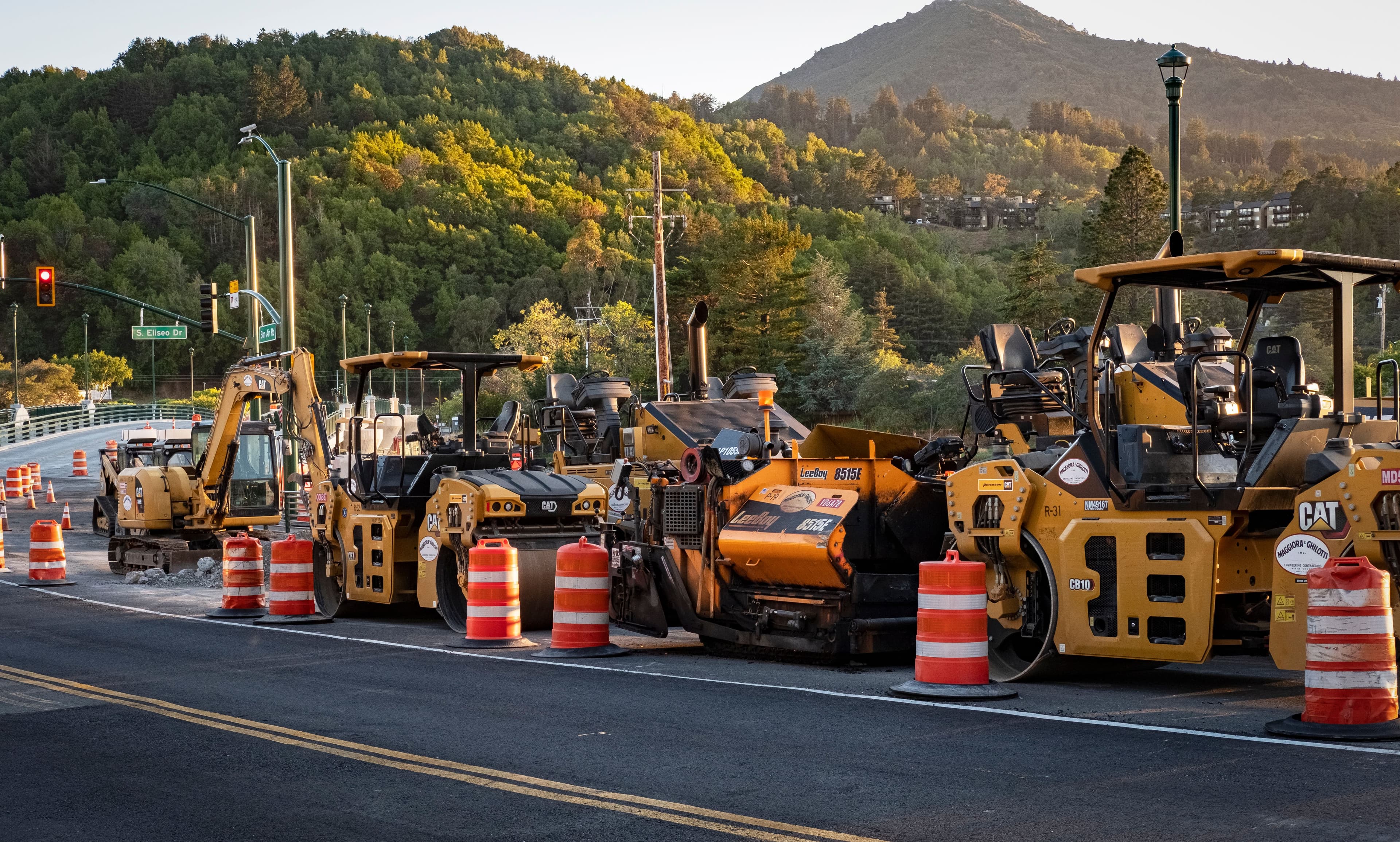 Yellow construction equipment and orange traffic barrels parked on a road near a wooded hillside.